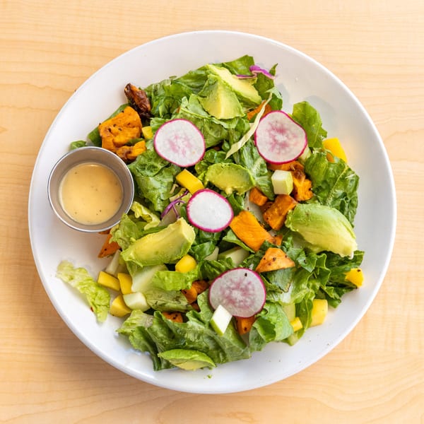 Overhead view of a vibrant salad with avocado, radishes, and roasted sweet potatoes, with dressing on the side.