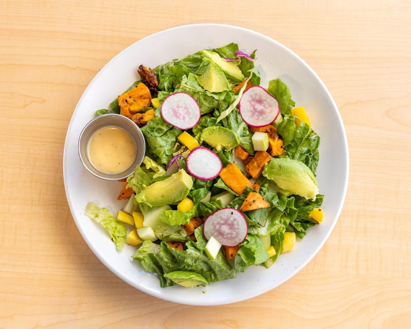 Overhead view of a vibrant salad with avocado, radishes, and roasted sweet potatoes, with dressing on the side.
