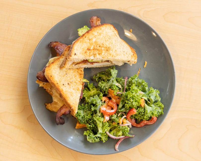 Grilled cheese sandwich with bacon and avocado, served with a side salad on a gray plate.