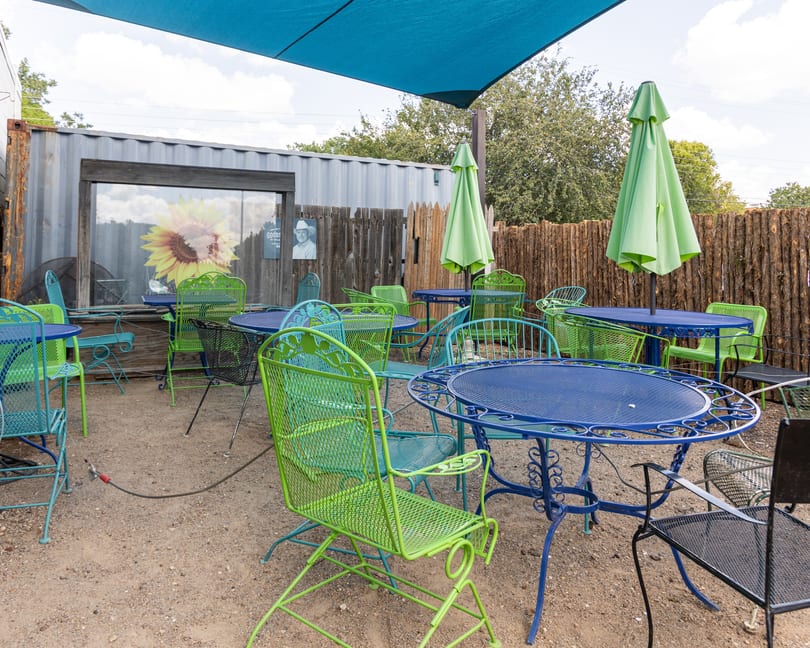 Outdoor patio with blue and green metal tables and chairs under a blue shade sail.