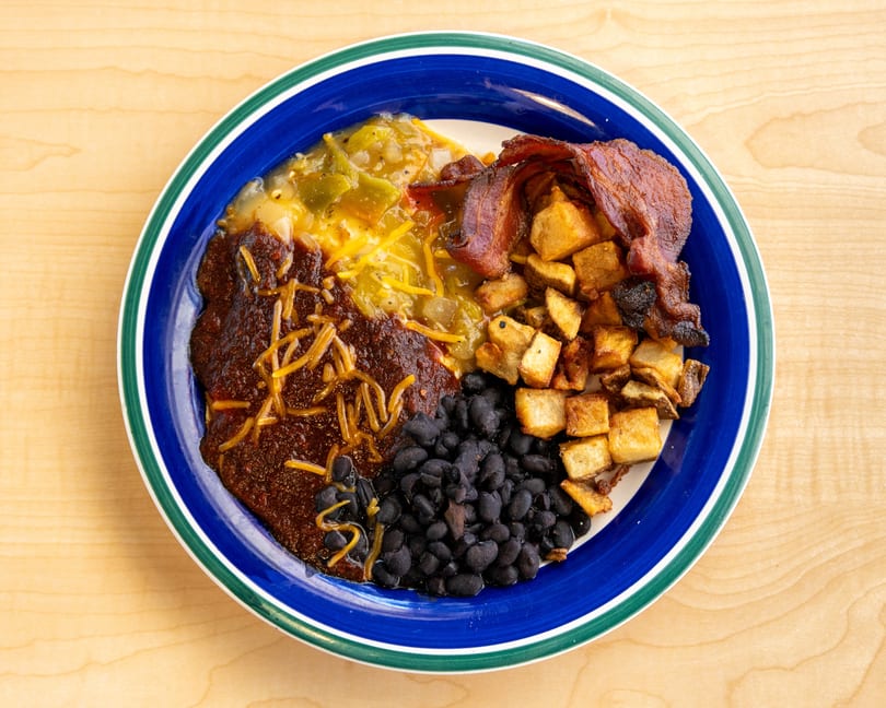 A blue plate with green and white trim holds a breakfast of chili, black beans, potatoes, and bacon.
