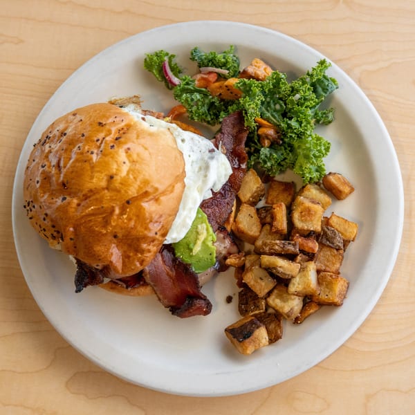 Breakfast sandwich with bacon, avocado, and fried egg, served with crispy potatoes and kale salad.
