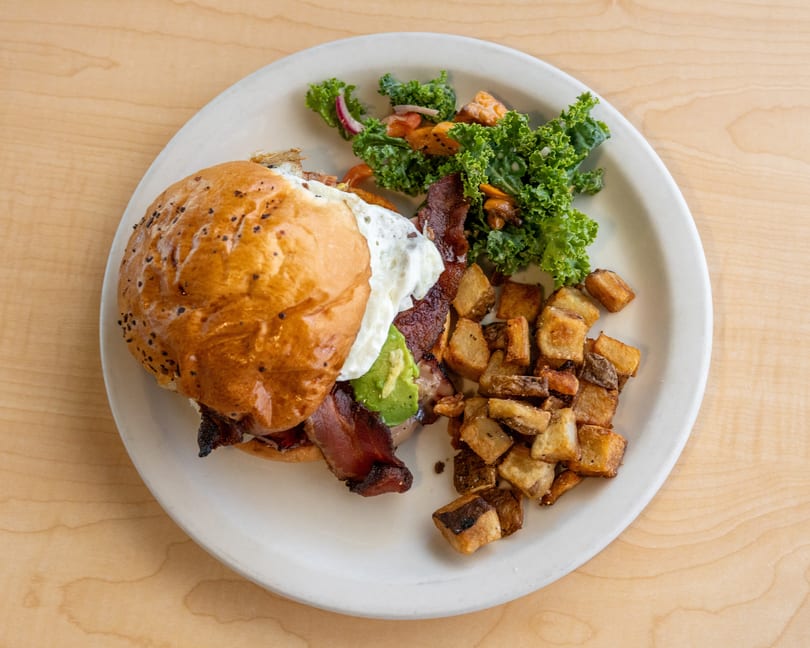 Breakfast sandwich with bacon, avocado, and fried egg, served with crispy potatoes and kale salad.