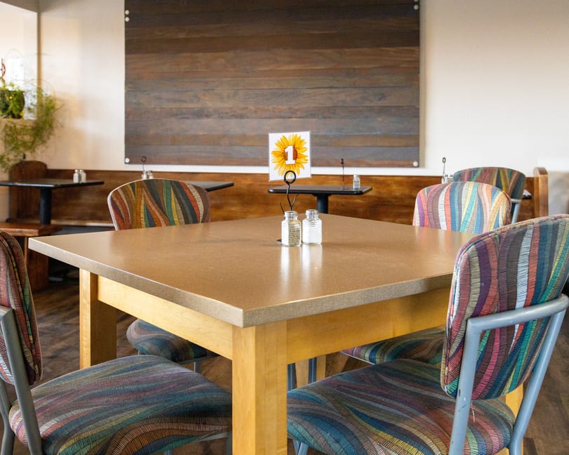 Restaurant dining area with a light brown table, colorful chairs, and a sunflower table number.