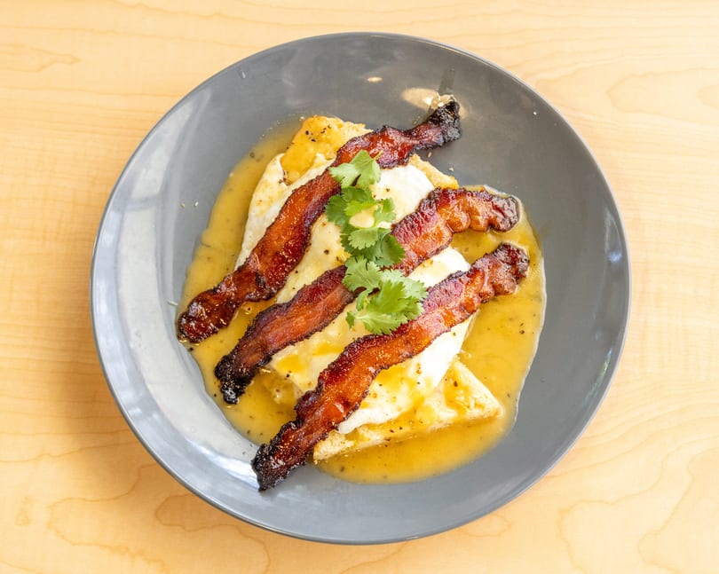 Crispy bacon strips and fried eggs with a yellow sauce and cilantro on a gray plate.