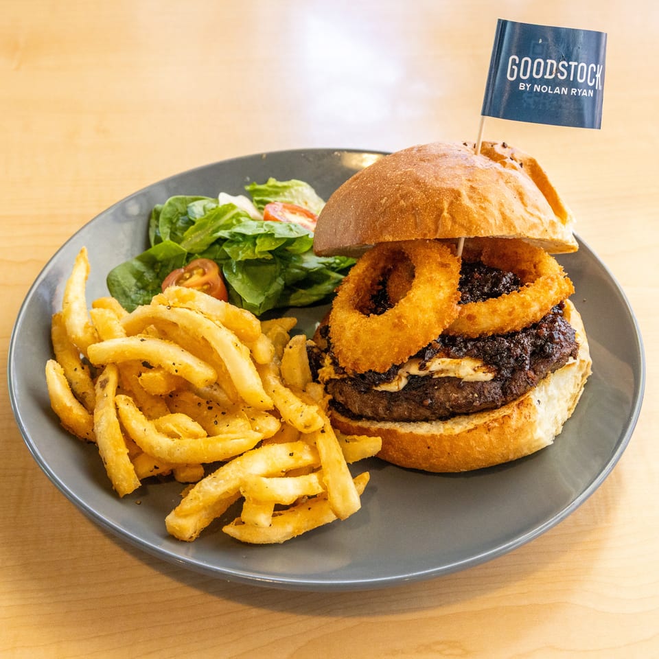 A Goodstock burger with onion rings, fries, and a side salad on a gray plate.