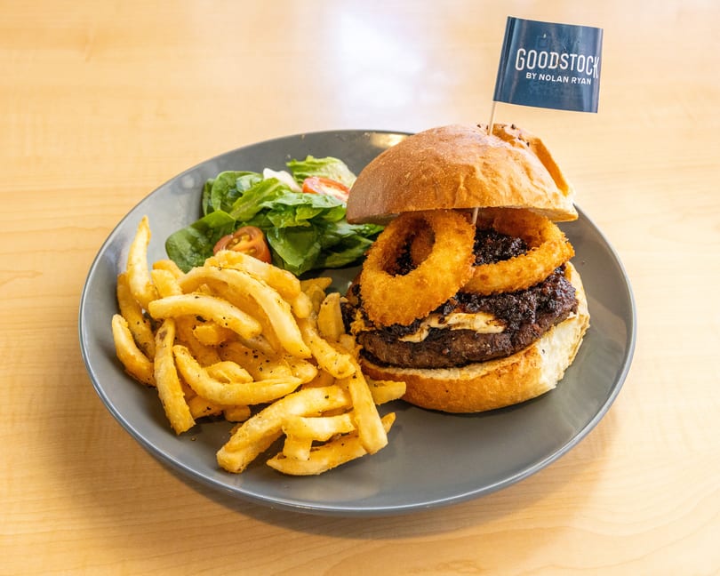 A Goodstock burger with onion rings, fries, and a side salad on a gray plate.