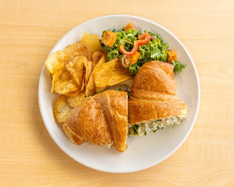 Chicken salad croissant sandwich with a side of chips and kale salad on a white plate.
