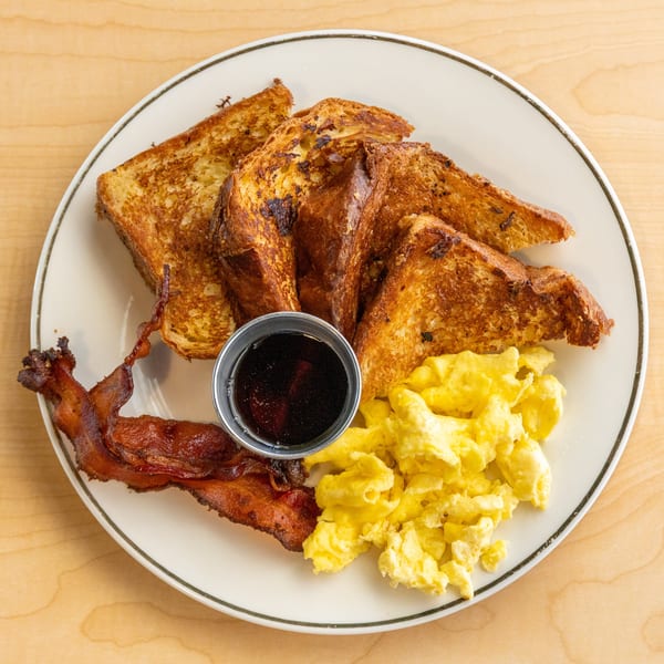 A breakfast plate with golden French toast, scrambled eggs, crispy bacon, and a side of syrup.