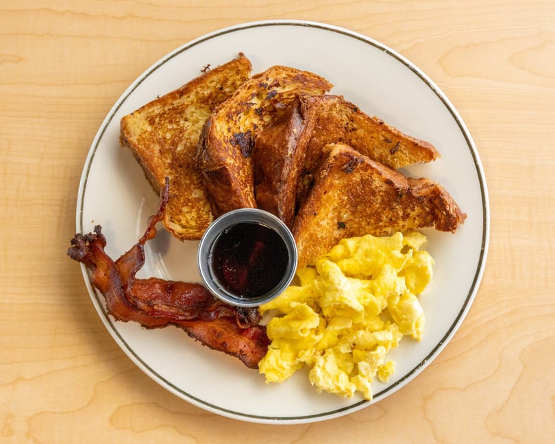 A breakfast plate with golden French toast, scrambled eggs, crispy bacon, and a side of syrup.
