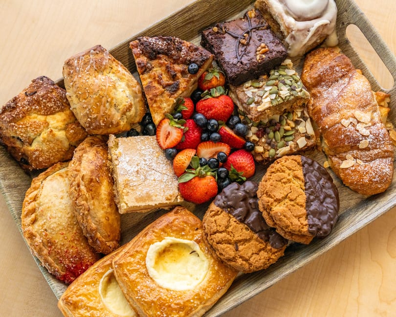 A wooden tray filled with assorted pastries, cookies, brownies, and fresh berries on a light wood surface.