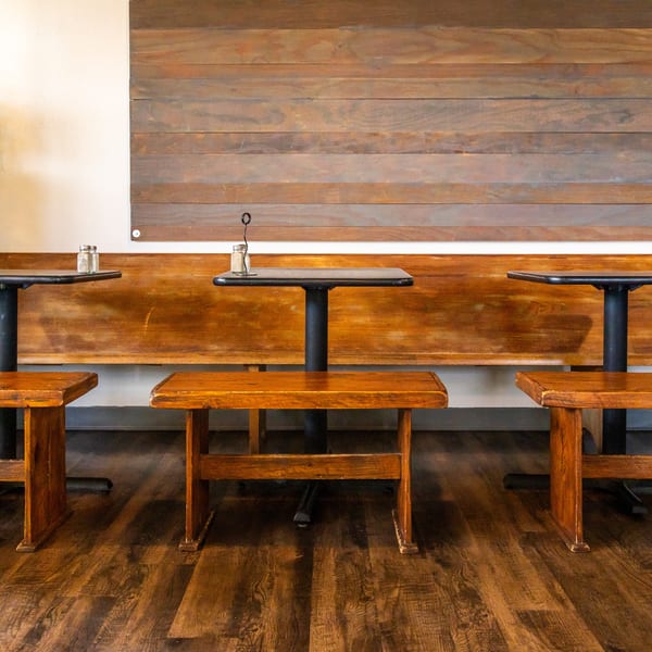 Three wooden tables with benches and a long wooden backrest against a white wall.