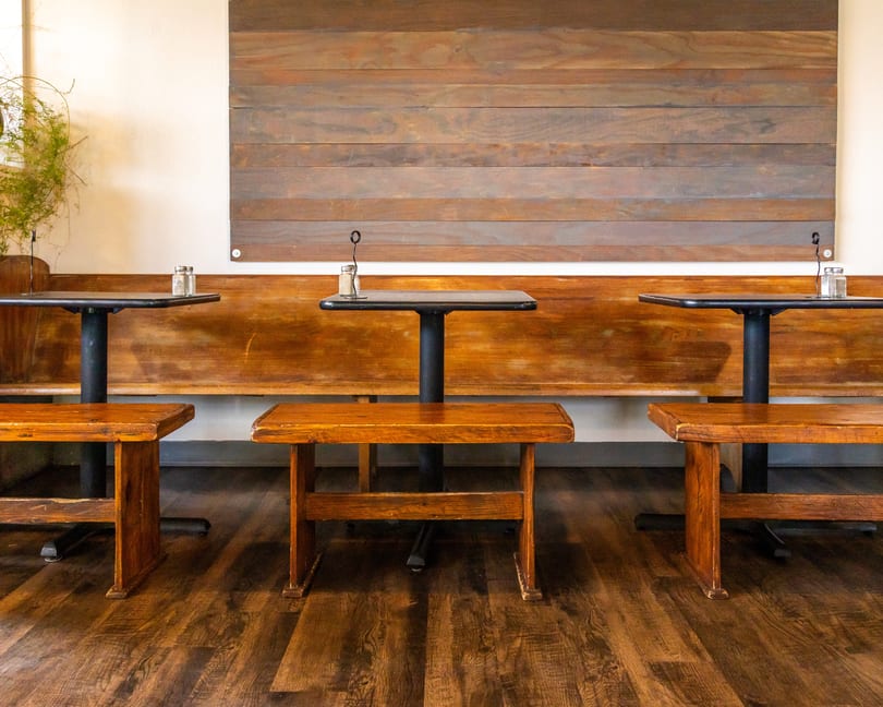 Three wooden tables with benches and a long wooden backrest against a white wall.