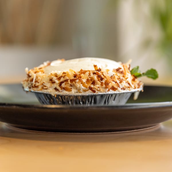 A small coconut cream pie in a foil tin, topped with toasted coconut flakes and a mint sprig.