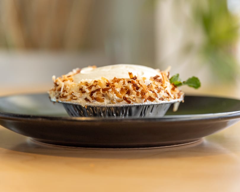 A small coconut cream pie in a foil tin, topped with toasted coconut flakes and a mint sprig.