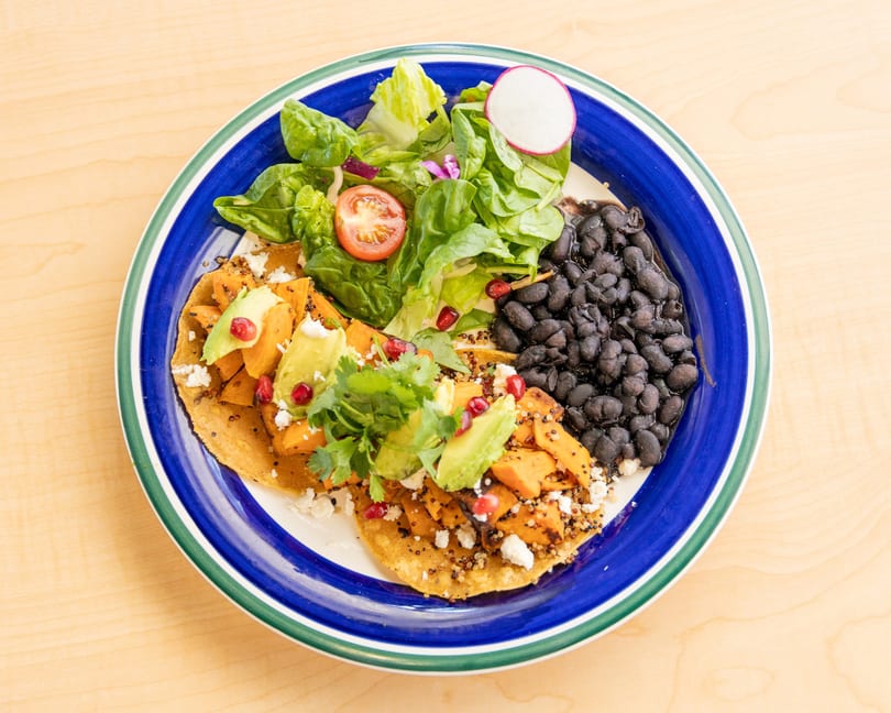 Sweet potato tostadas with avocado, cilantro, pomegranate, black beans, and a side salad on a blue plate.