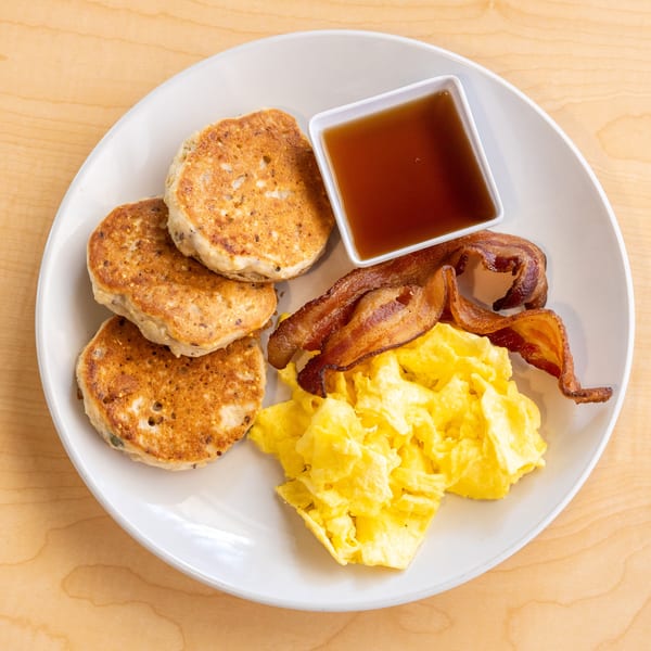 A breakfast plate with pancakes, scrambled eggs, crispy bacon, and a side of maple syrup.