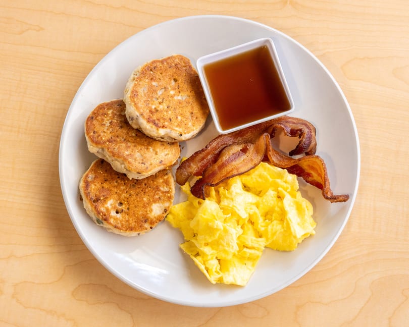 A breakfast plate with pancakes, scrambled eggs, crispy bacon, and a side of maple syrup.