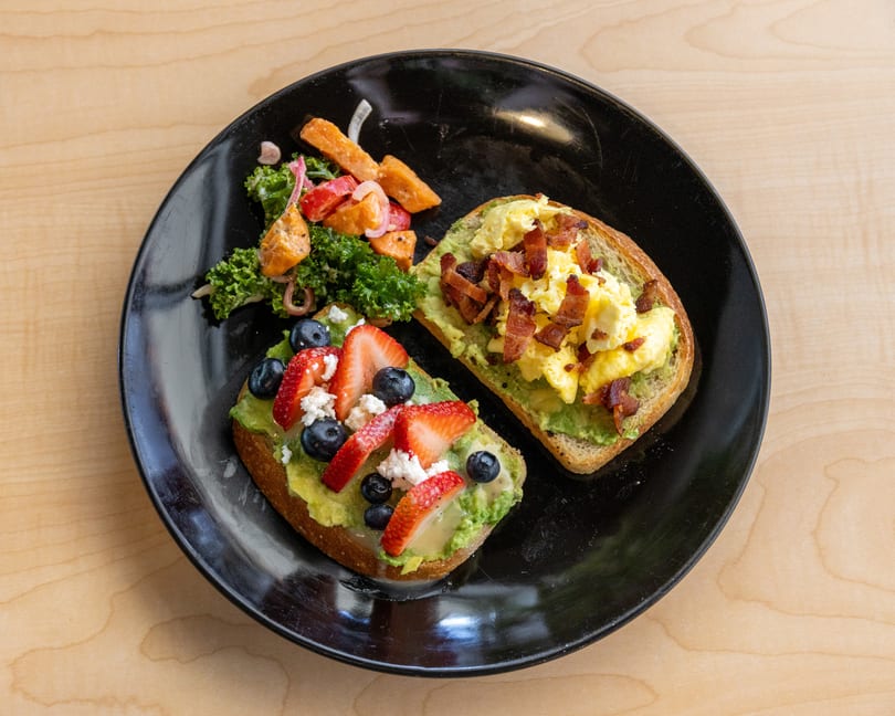 Two avocado toasts, one with scrambled eggs and bacon, the other with berries and feta, served with a side salad.