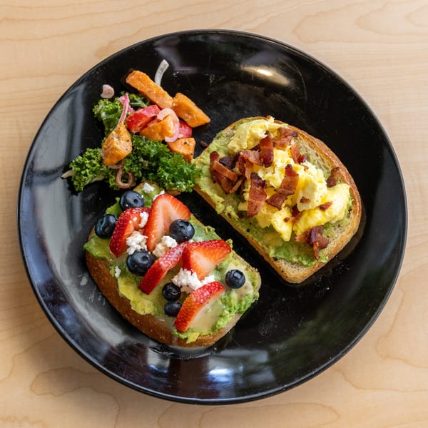 Two avocado toasts, one with strawberries and blueberries, the other with scrambled eggs and bacon, served with a side salad.