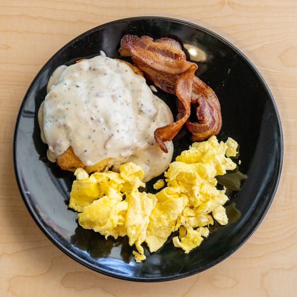 A black plate with biscuits and gravy, crispy bacon, and scrambled eggs on a wooden table.