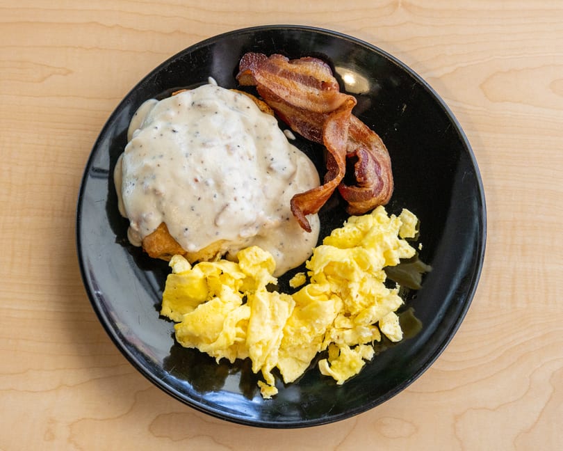 A black plate with biscuits and gravy, crispy bacon, and scrambled eggs on a wooden table.