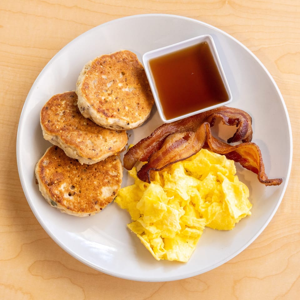 A breakfast plate with pancakes, scrambled eggs, crispy bacon, and a side of maple syrup.