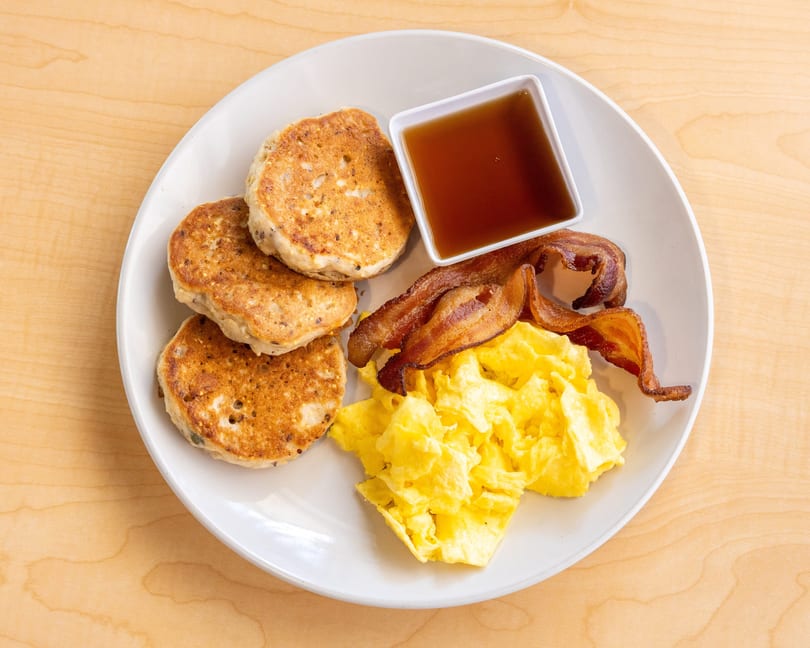 A breakfast plate with pancakes, scrambled eggs, crispy bacon, and a side of maple syrup.